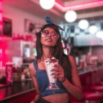 Young woman in fashionable attire enjoying a milkshake at a retro diner with vibrant lighting.