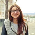 A young woman with glasses smiling outdoors in Budapest street scene, showcasing fashionable style.